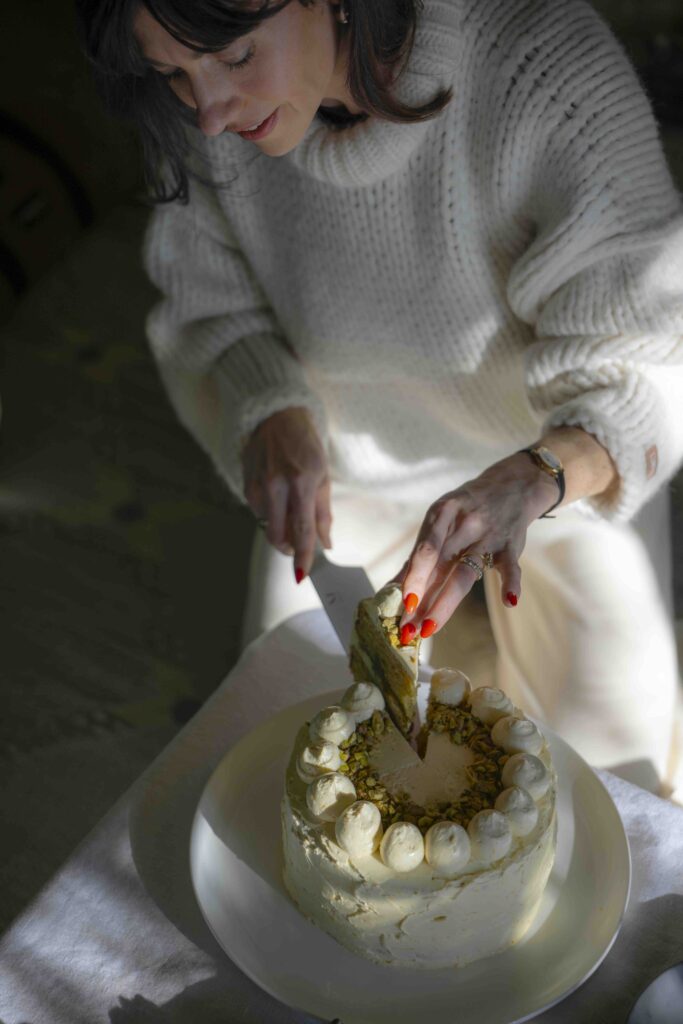 A woman is wearing a cream-colored sweater and pants, cutting into a ricotta olive oil cake