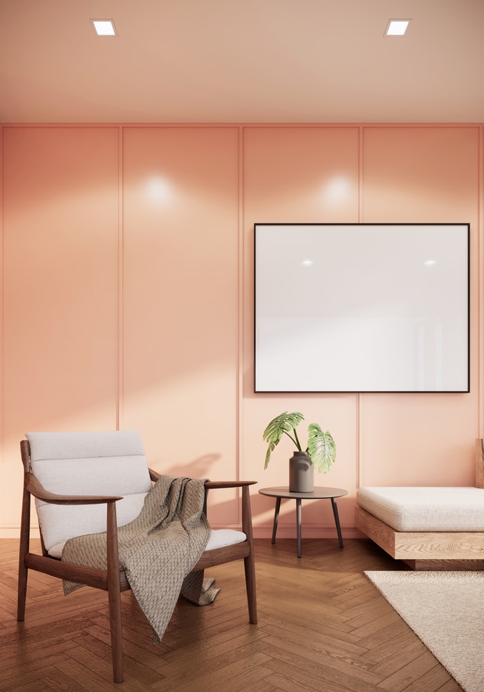 A modern living room with a mid-century style chair draped with a neutral throw, a white rug, wooden side table with a green plant and wooden floors. The wall behind is painted peach.
