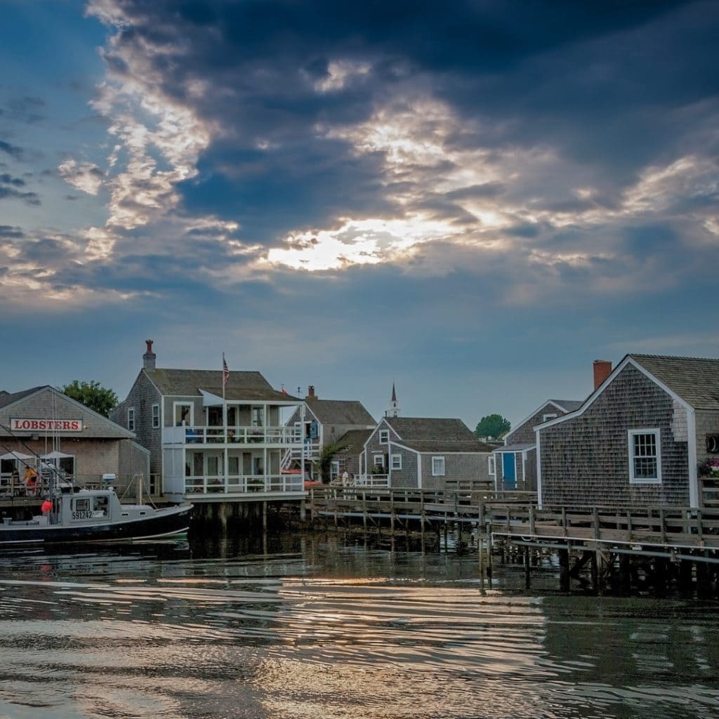 The shore of Nantucket with a storm brewing in the sky. There are several smaller, tan sided buildings, a lobster boat, and a small restaurant with a "lobsters" sign on the left.