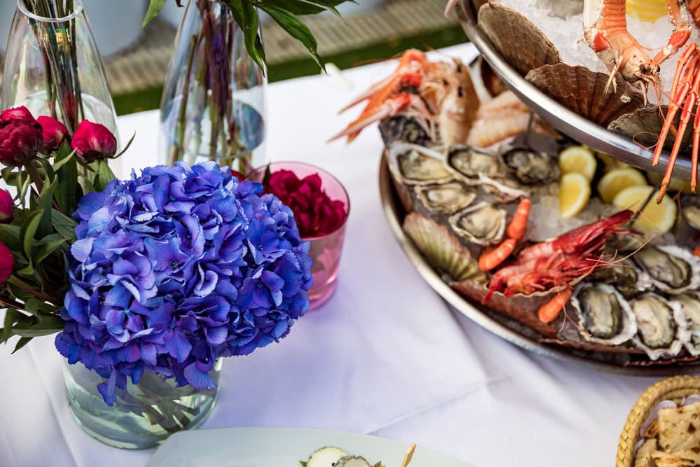 Oysters and shrimp on a tiered platter set on a white tablecloth next to purple hydrangea for a dark and stormy cocktail and themed party.