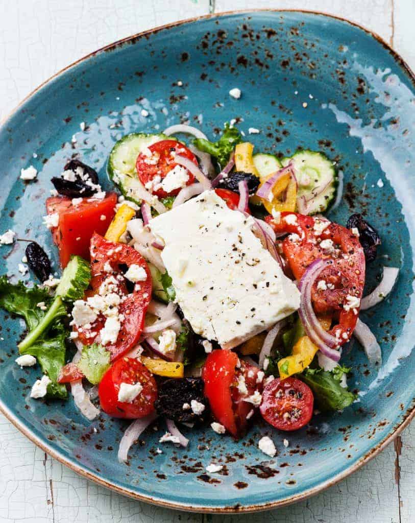 Salad with feta cheese, tomatoes, red onions, black olives on a blue plate.