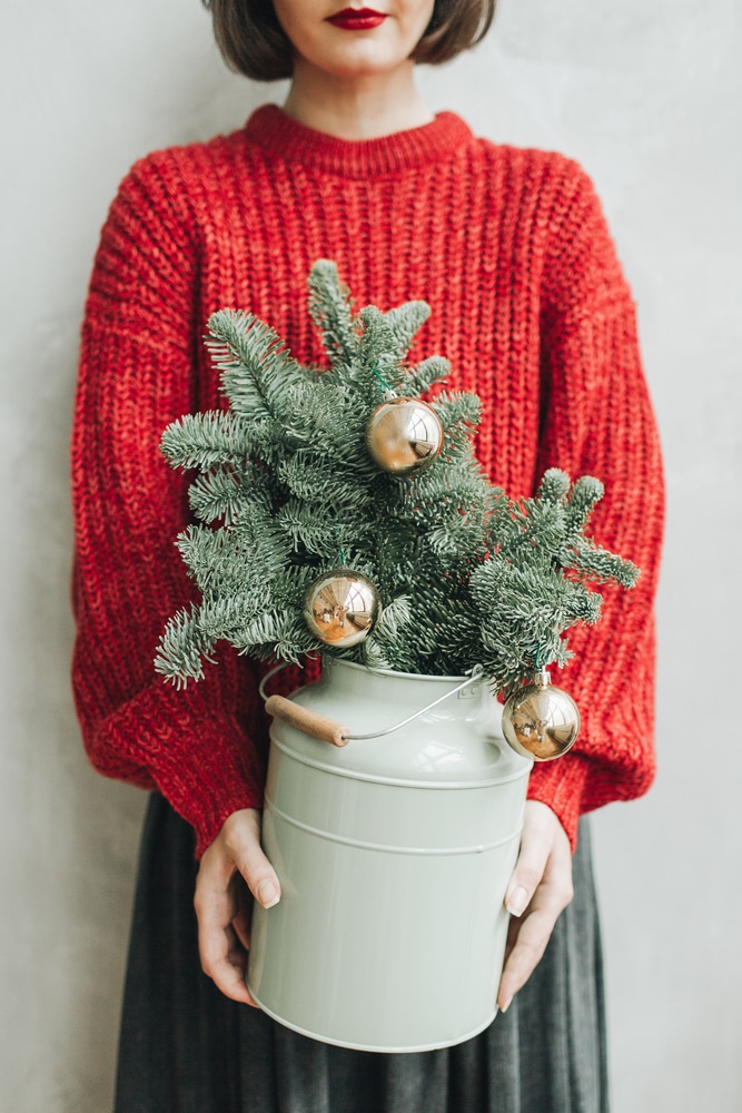 Young pretty woman with red knitted sweater and grey skirt holding in her hands vintage mint watering pot with Christmas fir branches decorated with gold balls. 