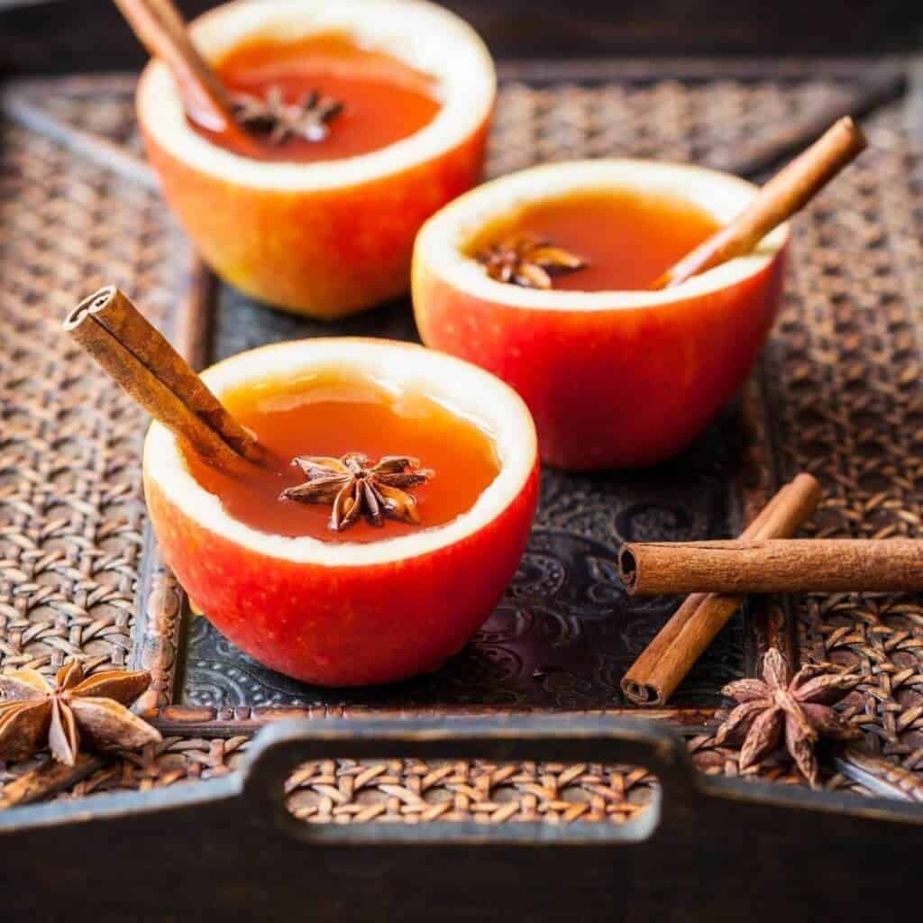 Hollowed out apples filled with hot apple cider, a cinnamon stick and star anise on a darker wicker tray with a mental part in the middle. There are cinnamon sticks crossed on the bottom right as well.