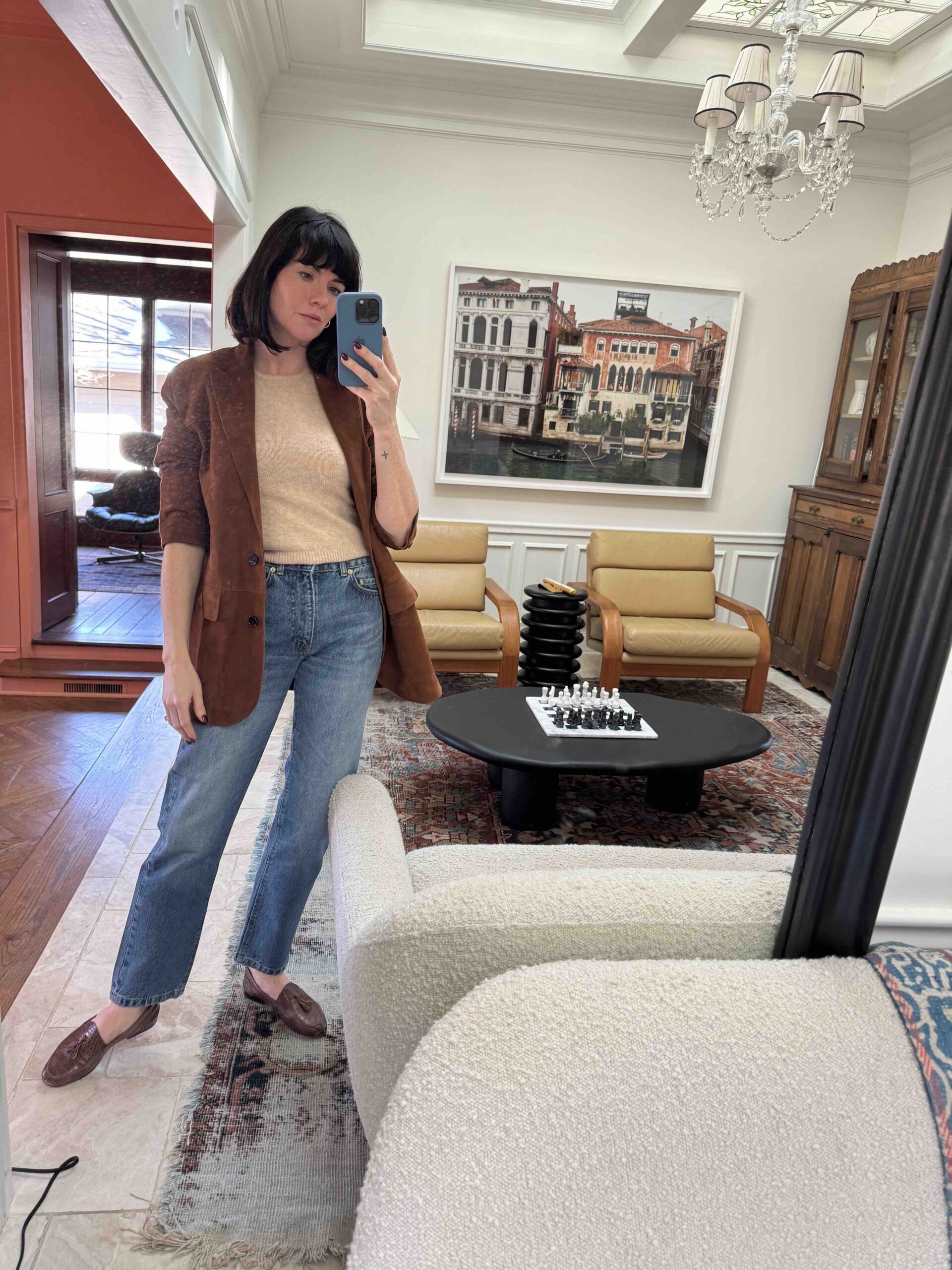 A woman wears blue jeans, a beige sweater tank, a brown blazer, and brown loafers