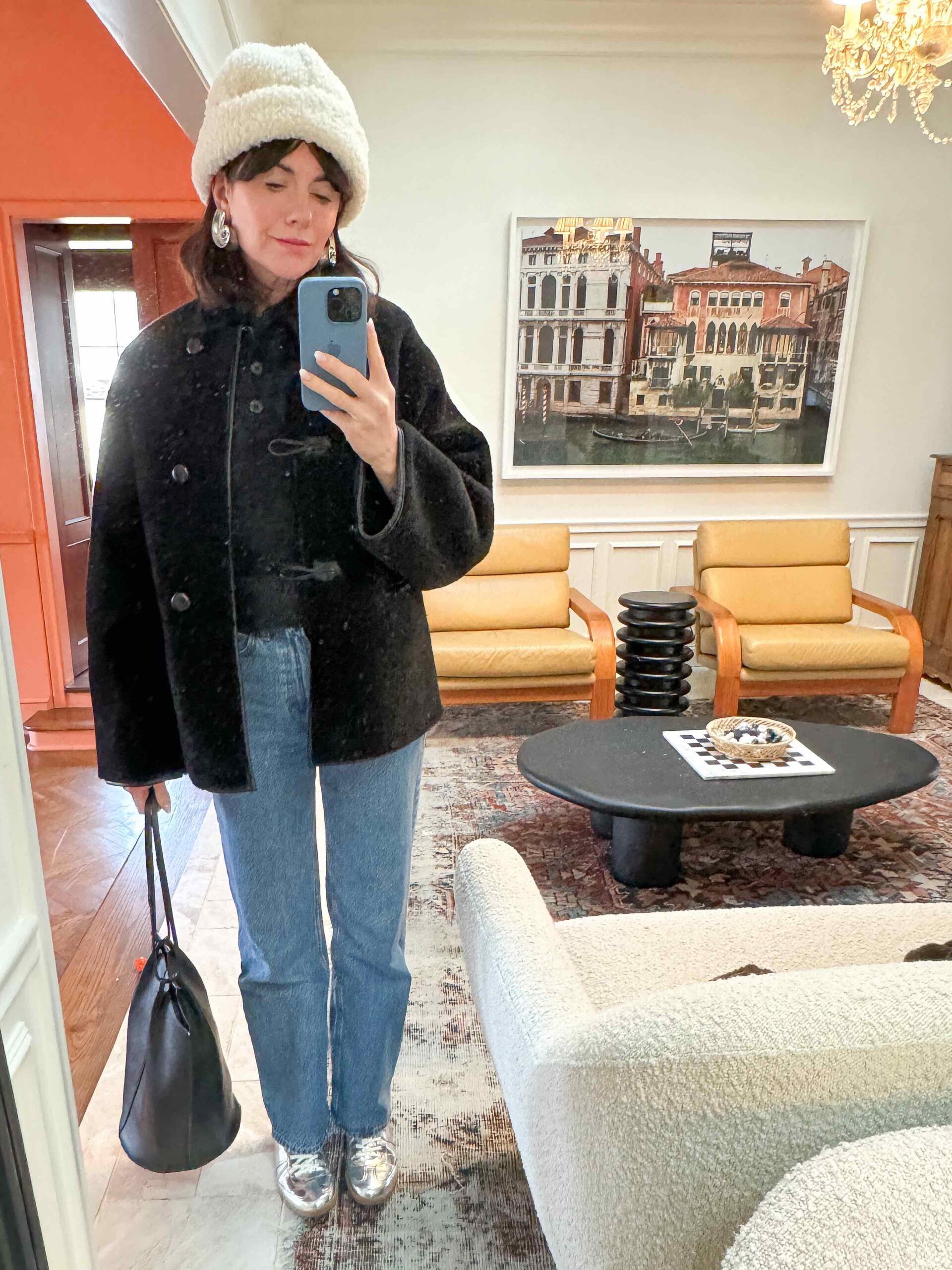 A woman wears a black polo sweater, black shearling coat, blue jeans, silver sneakers, a white hat, silver earrings, and a black handbag