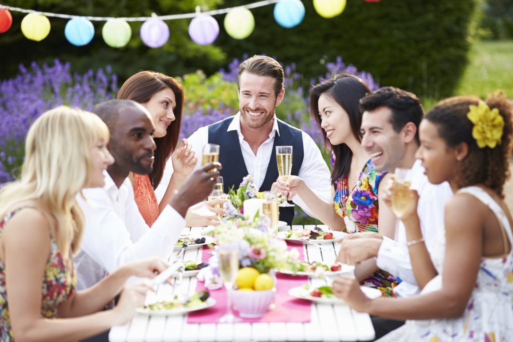 Group Of ethnically diverse Friends Enjoying Outdoor Dinner Party. It's a white picnic table with purple and green foliage in the background.