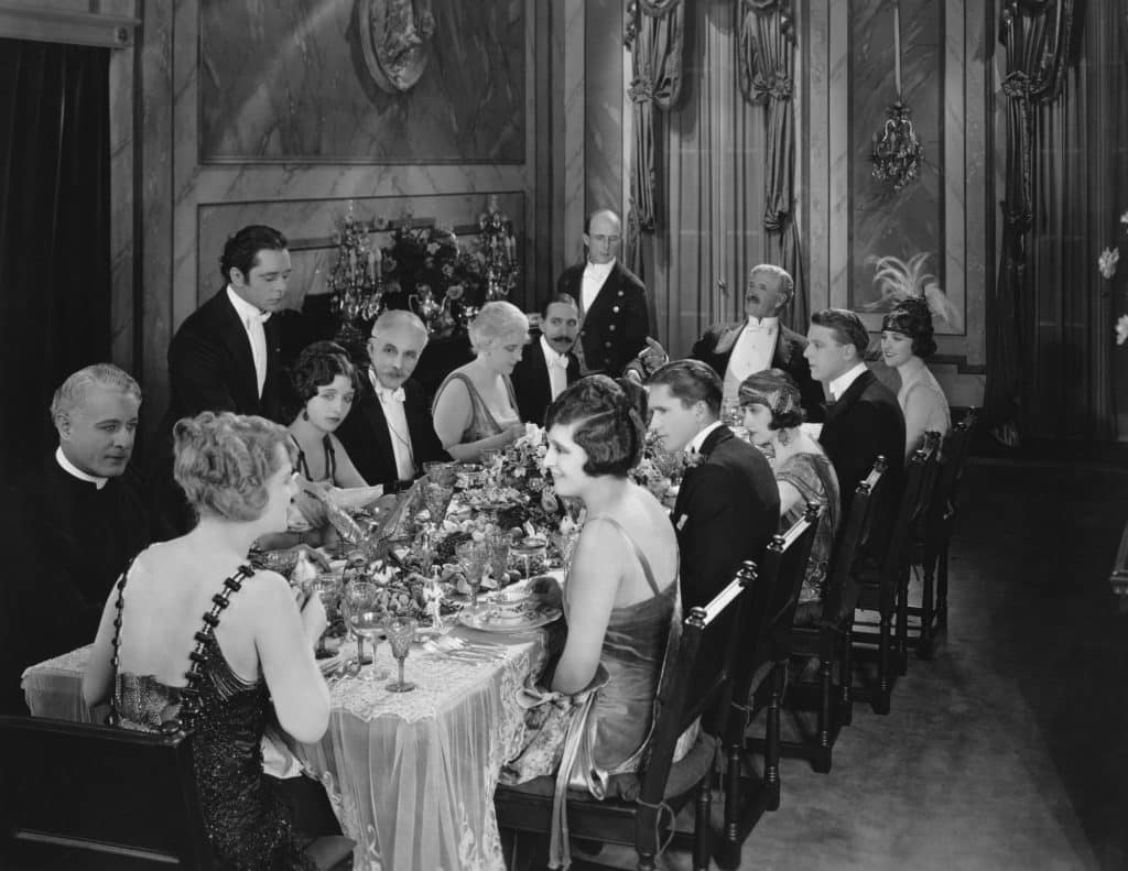 Black and white photo from what looks like it may be the 1920's of a group of 12 people having dinner and two butlers serving them.