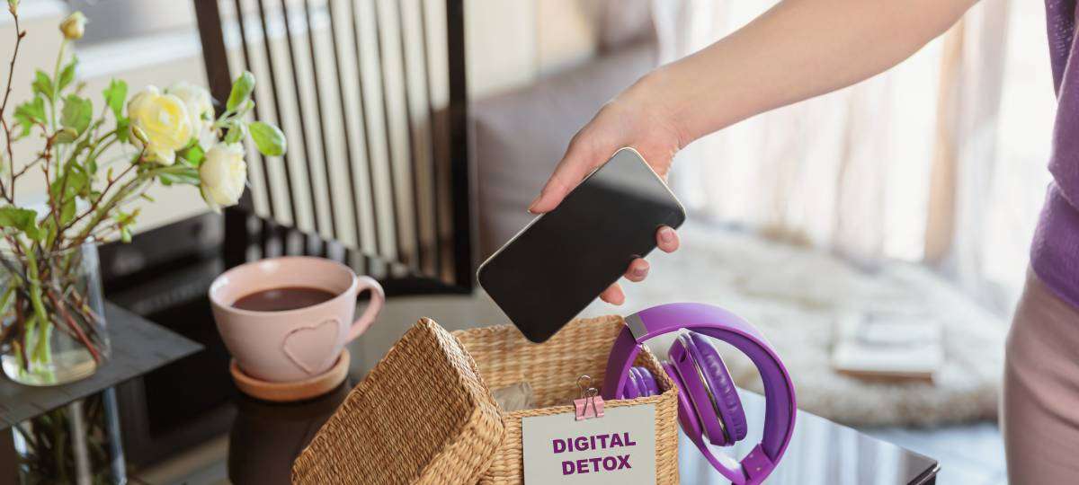 Close-up of an arm putting a smartphone in a basket that says "Digital Detox." The box also has a pair of headphones inside.