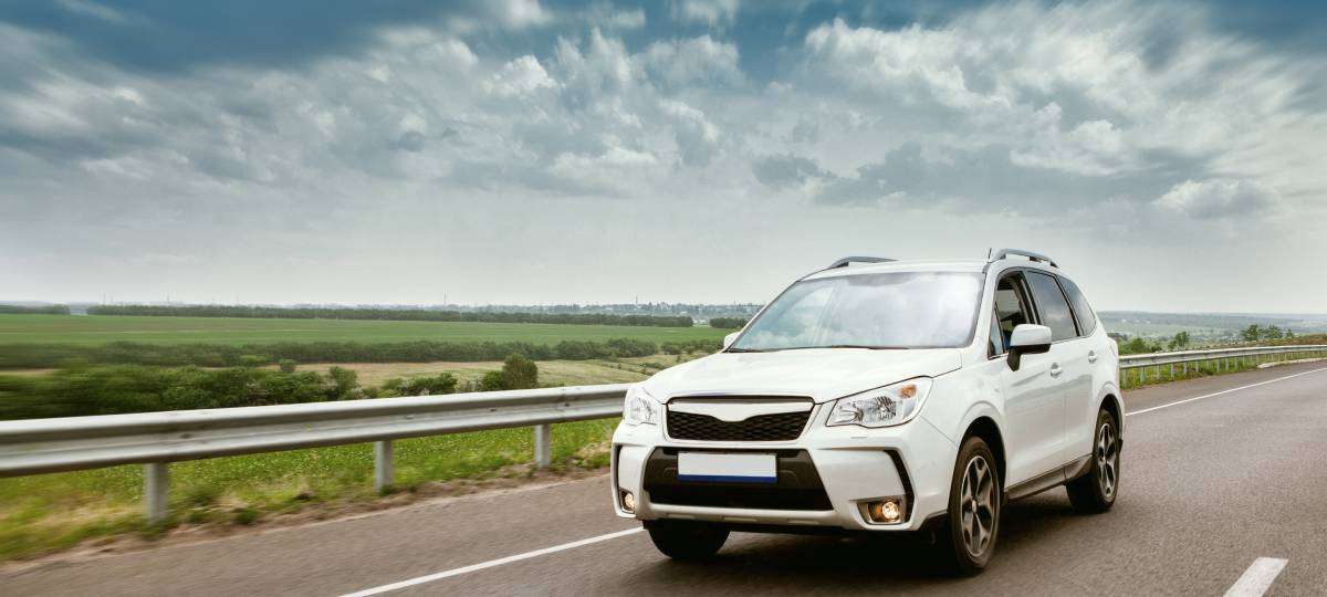 A small white SUV drives alone down a black asphalt road with white lines. The countryside appears in the background.