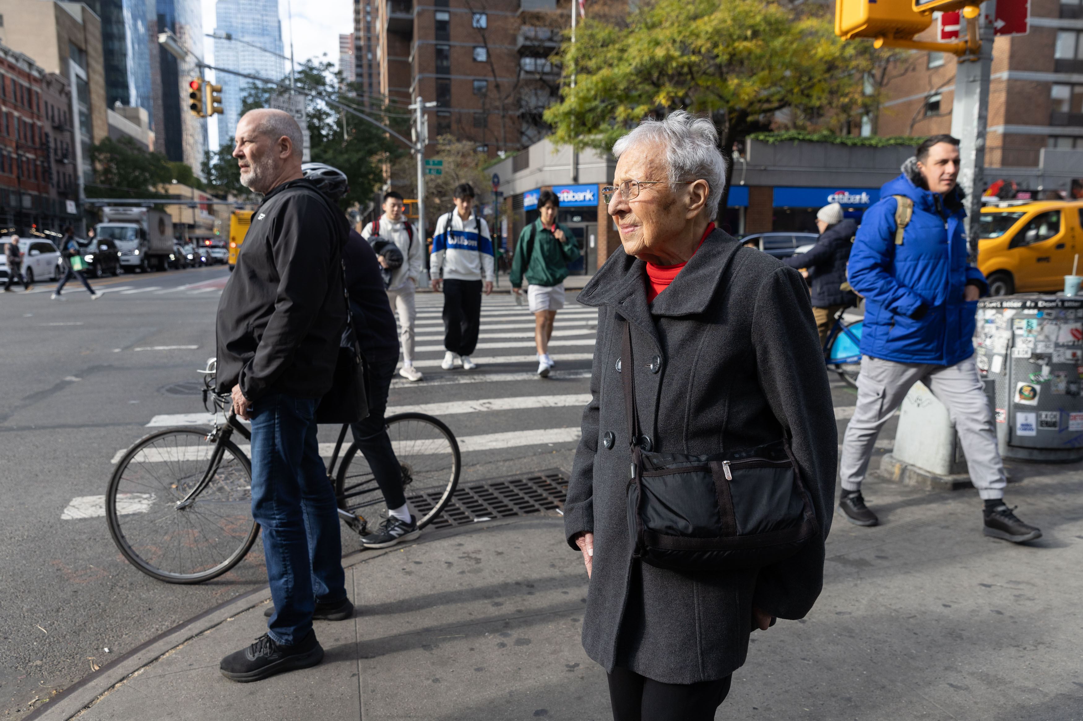 A photograph of a senior woman standing on the corner of a busy block in the Upper West Side of New York on a cold, but sunny day.