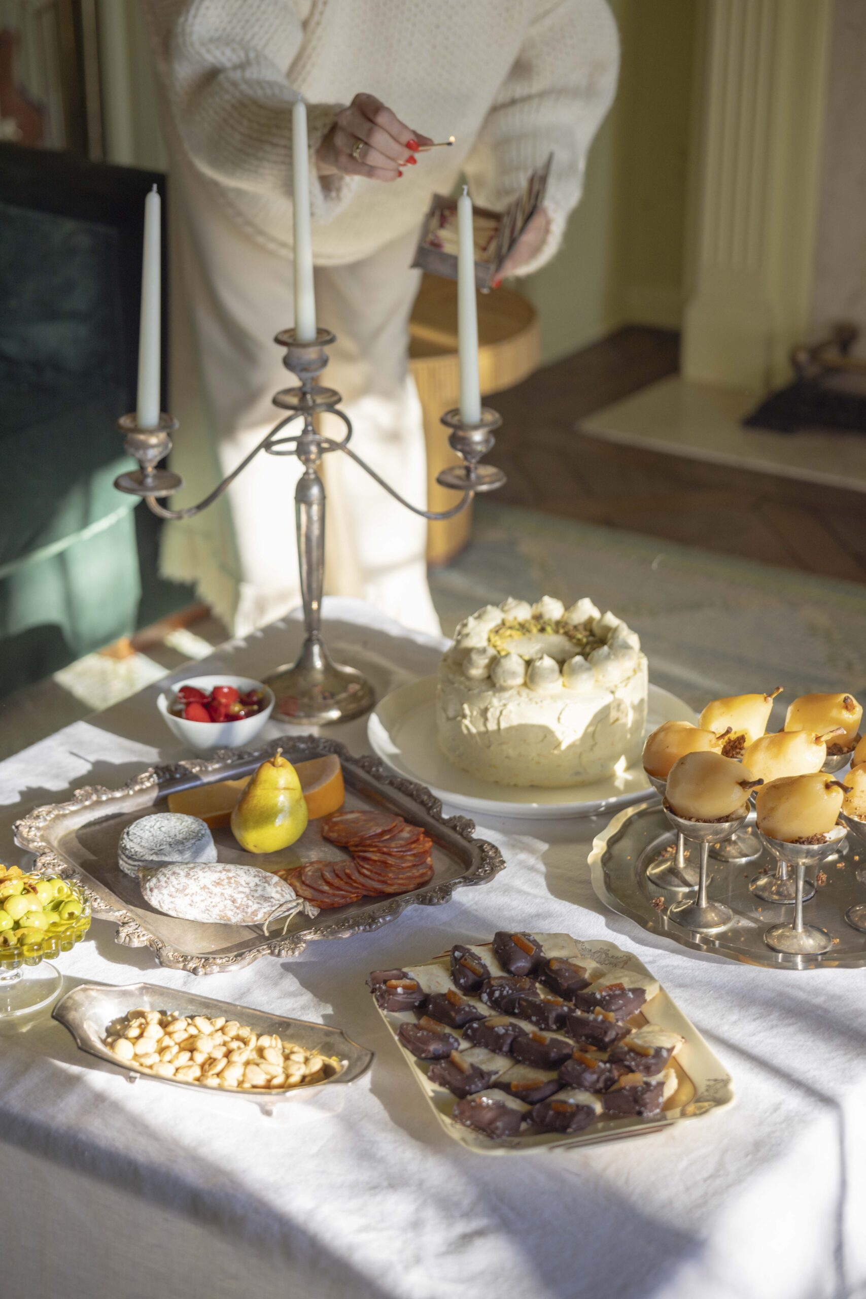 A woman lights tapered candles while a coffee table is set for an afternoon dinner party. Serving trays hold shortbread cookies, ricotta olive oil cake, poached pears, and charcuterie