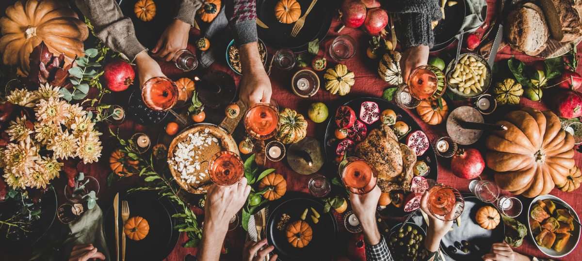 A top-down view of a table packed with classic Thanksgiving dishes and several hands reaching out to clink wine glasses.