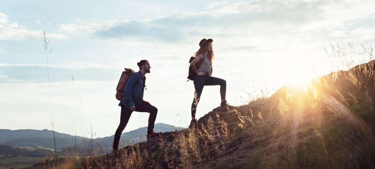 Two people hiking up a hill during the day while wearing backpacks. The person leading the hike is wearing a hat.