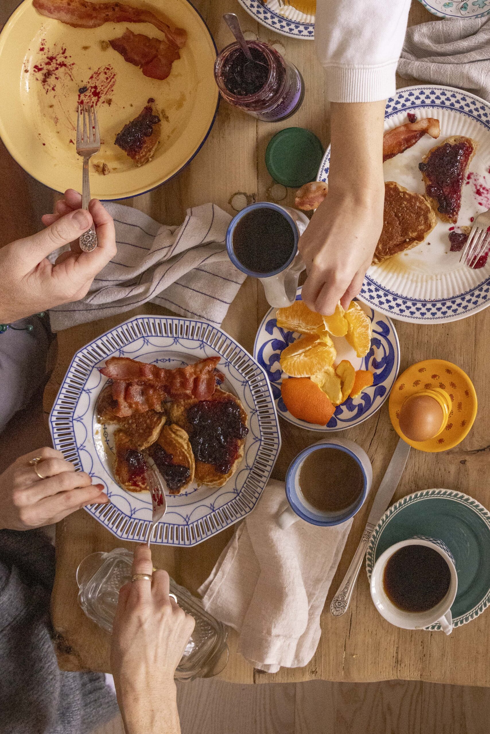 A wooden dining table is full of plates and coffee cups as friends enjoy buttermilk bran pancakes, bacon, and oranges for brunch