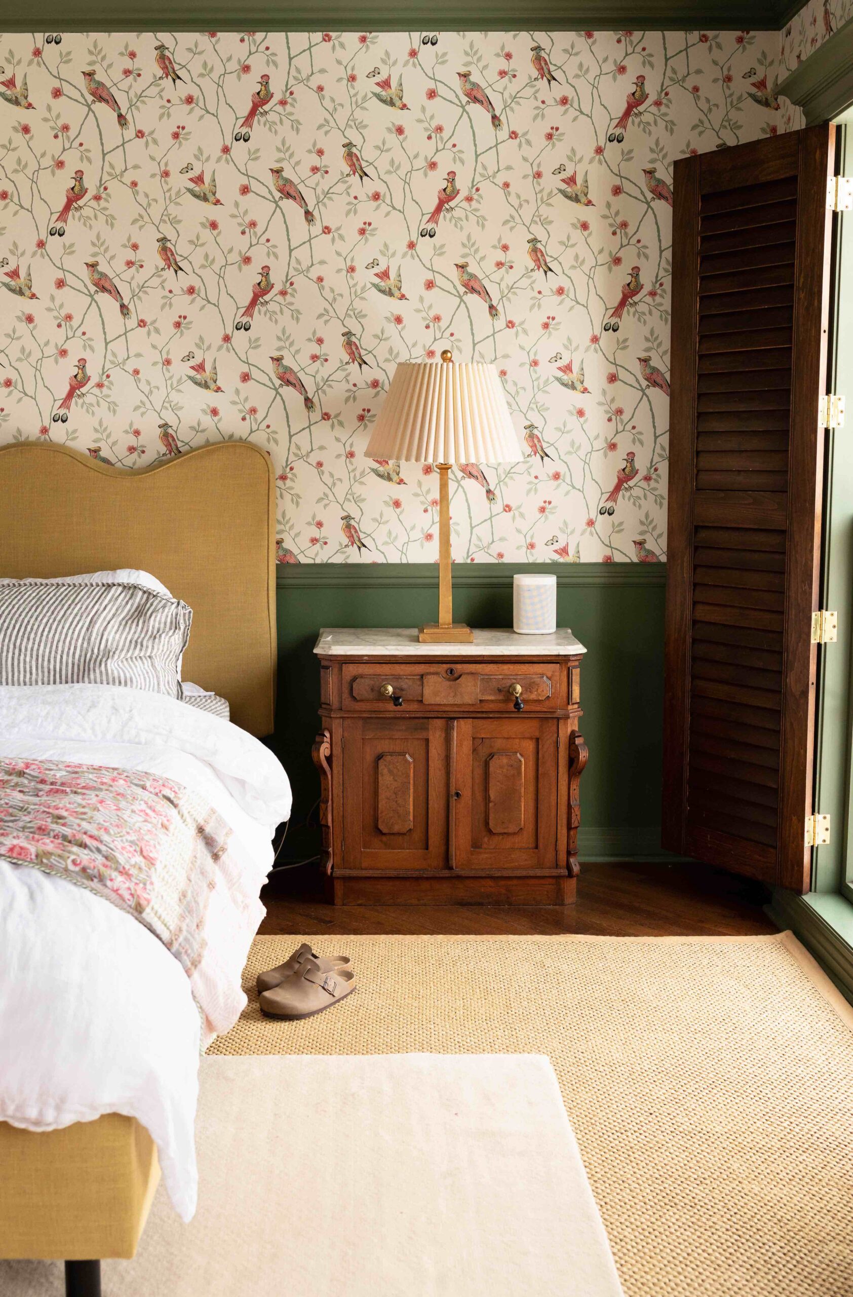 A primary bedroom features pink and green bird wallpaper, a scalloped headboard, layered rugs, interior shutters, and an antique wood nightstand with a white marble top