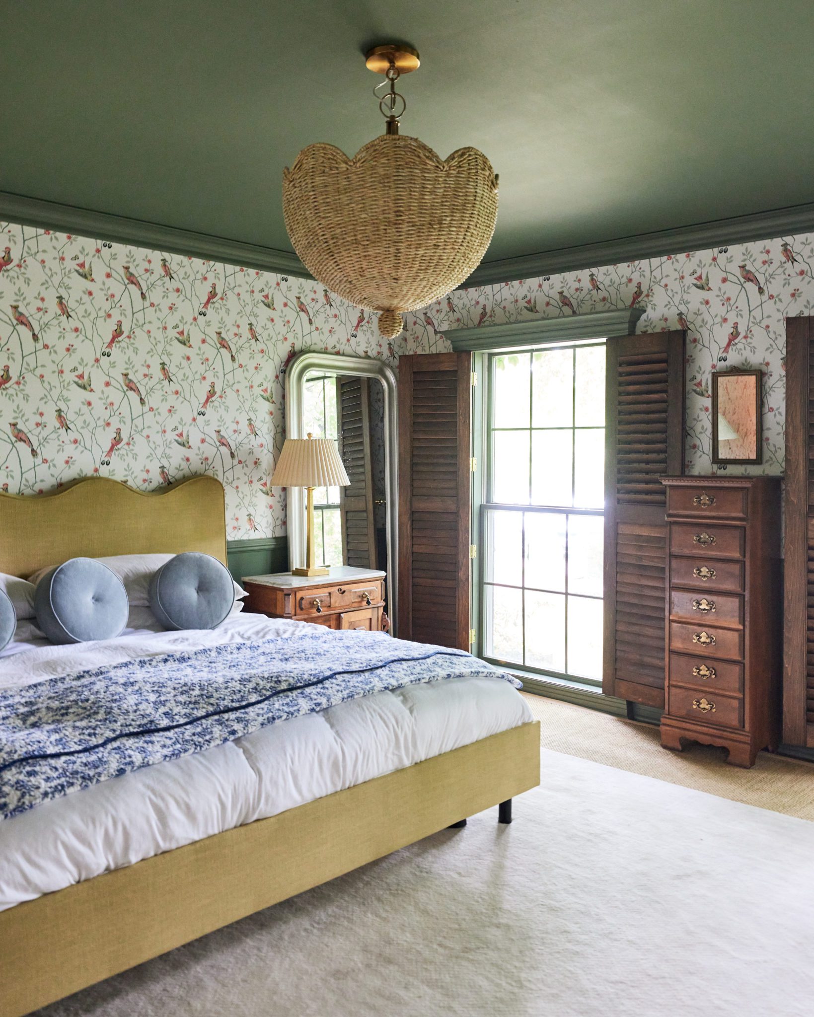 A main bedroom features a cream rug, an upholstered bed frame with a scalloped headboard, an antique dresser and nightstand, and walls covered in green and pink bird wallpaper